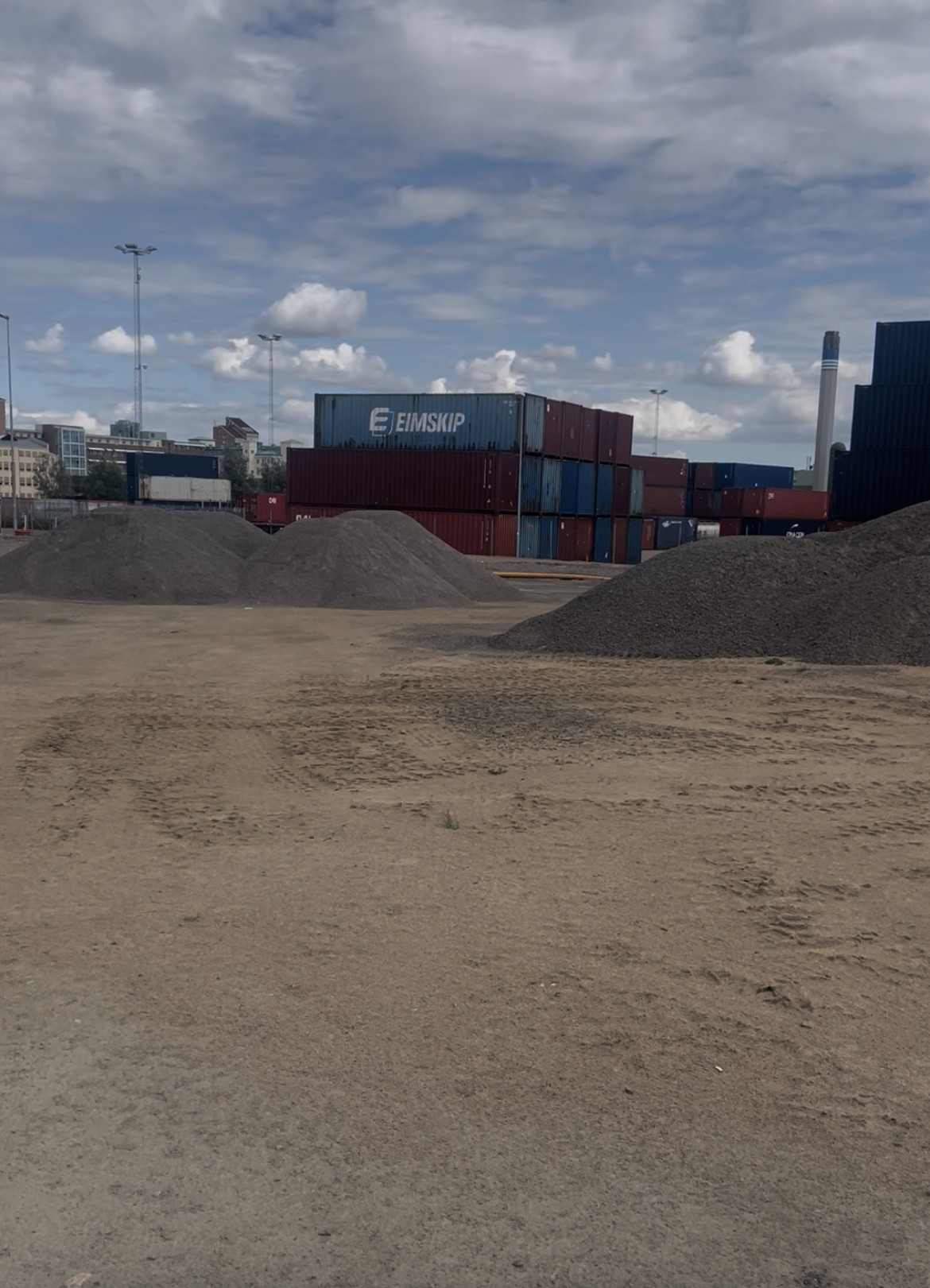 Grusplan och jordhögar på hamnområdet i Oceanhamnen med staplade lastcontainrar och stadens bebyggelse i bakgrunden.